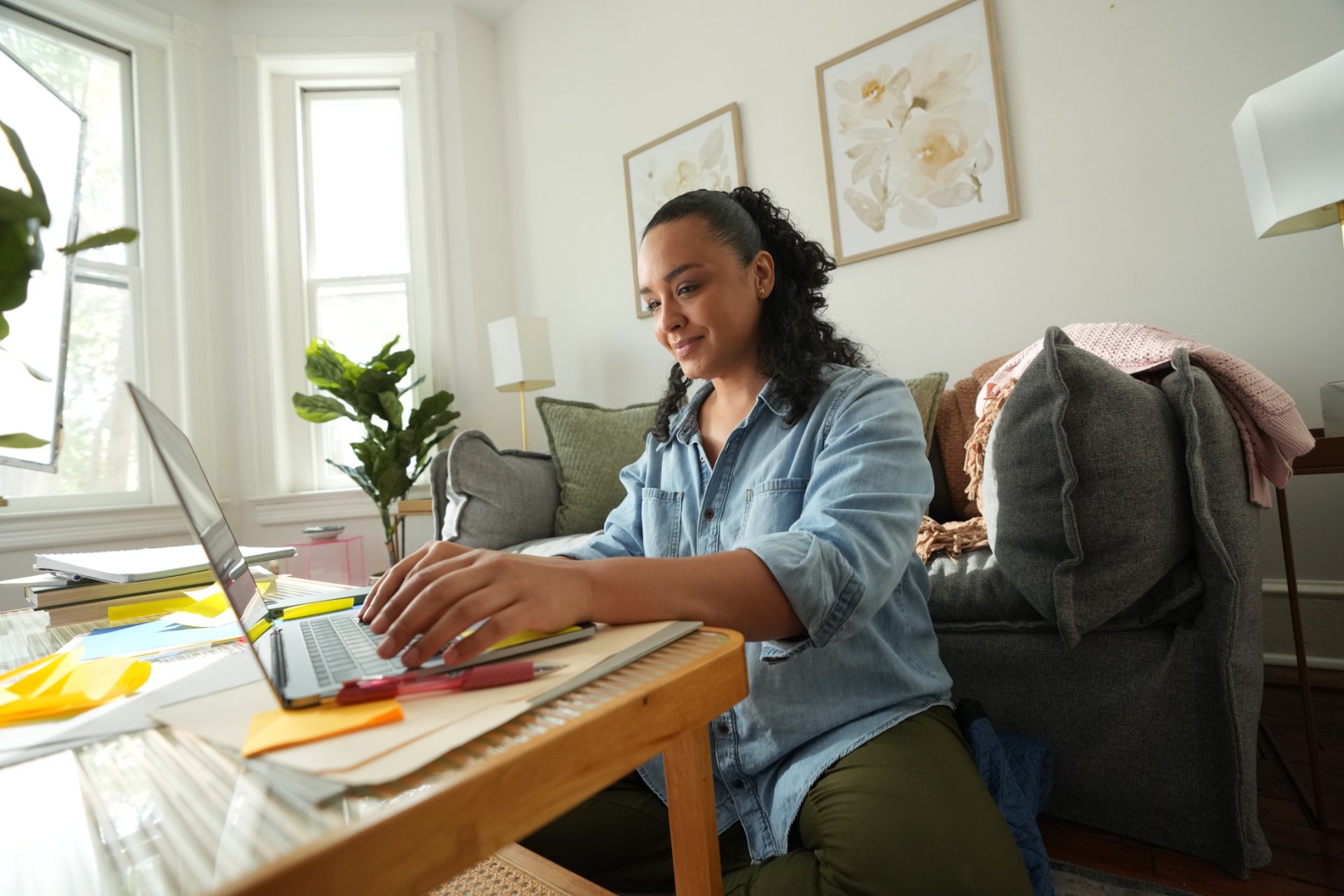 A woman sits at a coffee table in a bright living room using a laptop. Papers, notebooks, sticky notes, and pens are spread across the table as she works. A couch, houseplants, and framed floral artwork are visible in the background